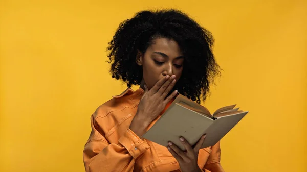 African american woman covering mouth while reading book isolated on yellow