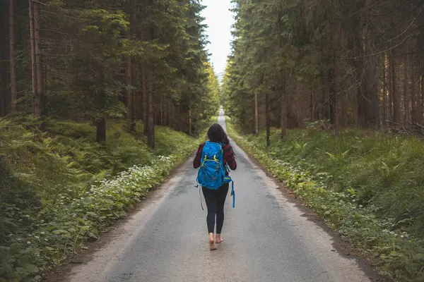 Back view of a woman with a blue backpack walking barefoot down a peaceful forest road, surrounded by lush greenery and tall trees, symbolizing adventure and tranquility in nature.