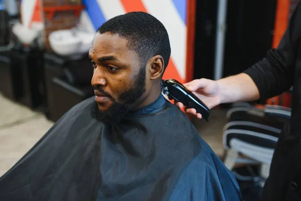 Portrait of young black man being trimmed with professional electric clipper machine in barbershop.Male beauty treatment concept. Young African guy getting new haircut in barber salon